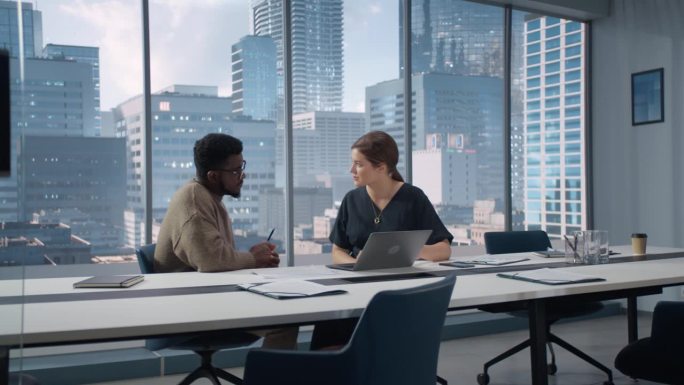 两个多民族同事在明亮的现代摩天大楼办公室里讨论。黑人创意总监和女性项目经理谈论笔记本电脑上的数据。