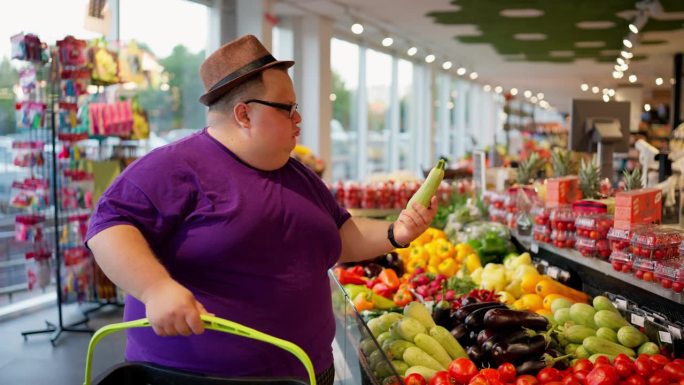在一家大型超市里，一个穿着紫色t恤、戴着眼镜、戴着棕色帽子的超重男子走过蔬菜柜台，挑选西红柿和西葫芦