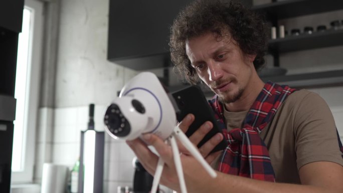 白人男子拿着家里的安全监控摄像头和手机试图安装一款连接安装和安装闭路电视安全视频监控摄像头监控系统慢