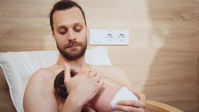 在医院的产后部，可爱的新生男婴在父亲的乳房上照顾他的宝宝，做皮肤对皮肤的特写。父亲坐在医院的扶手椅上