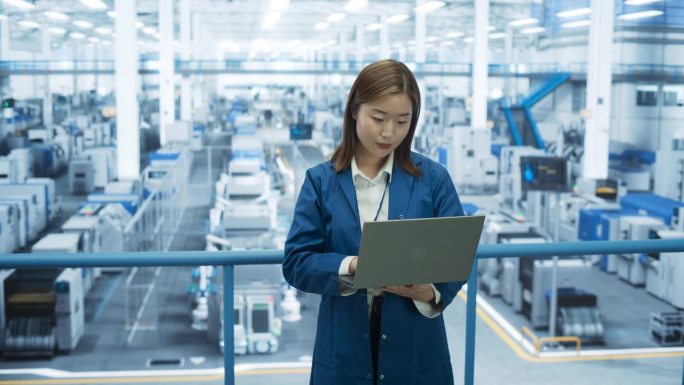年轻的亚洲女工程师站在工厂里使用笔记本电脑。机器在工作，专家通过在线系统监控进度