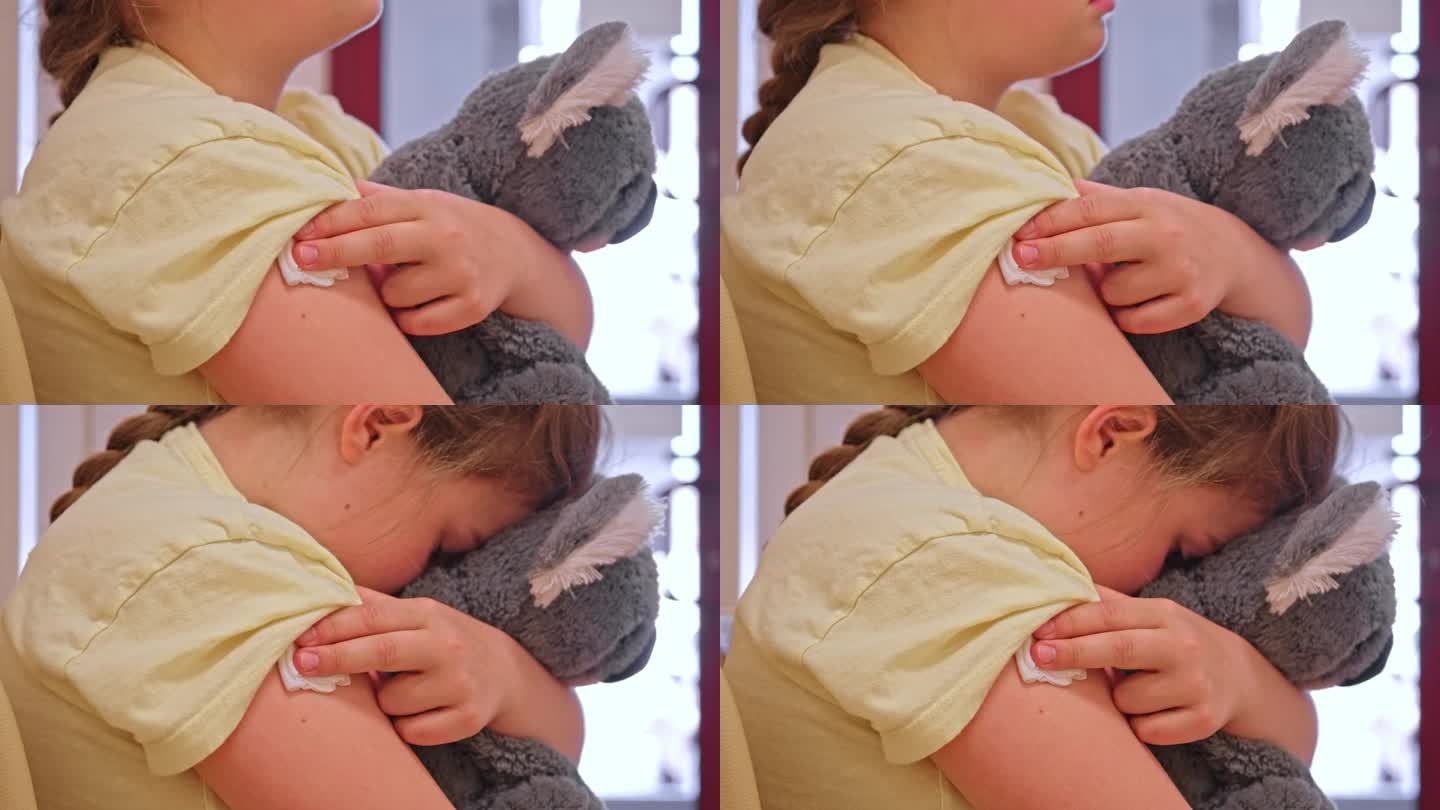 注射疫苗后，白人女孩抱着泰迪熊坐在诊所候诊室里，用纱布垫压着针头止血