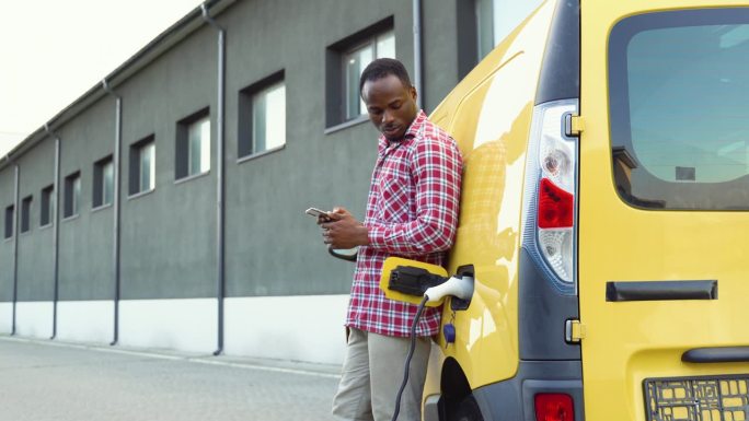 黑人男子手持智能手机在电动汽车充电站给卡车充电