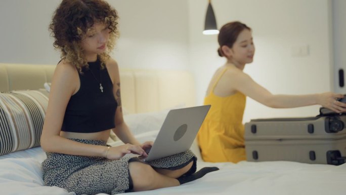 年轻女子把衣服装进行李箱，她的朋友盯着笔记本电脑屏幕计划度假。
