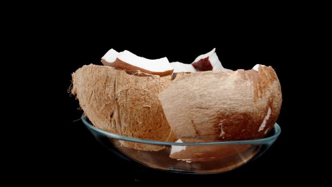 一个棕色的椰子碎成碎片，白色的果肉放在一个玻璃盘子里。它在一个孤立的背景上循环旋转。