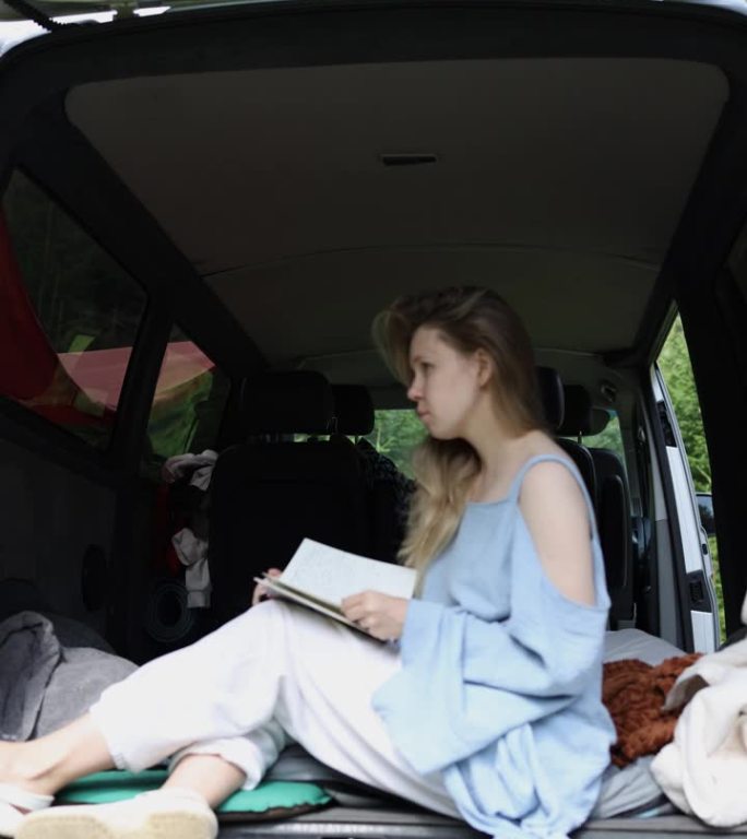 一个女孩在一辆小型房车的后备箱里看书。女孩合上笔记本，站起身来，带着一只白狗离开了画框。垂直视频