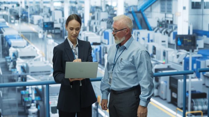 经验丰富的男女工程师站在现代工业工厂的平台上，使用笔记本电脑，讨论人工智能机器的现代电子产品生产