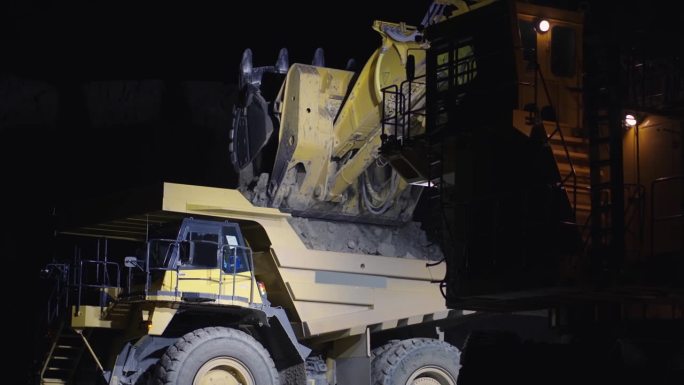 夜间在露天矿坑作业的挖掘机。画面中，挖掘机翻动着装有矿石的黄色铲斗进入自卸车，慢动作