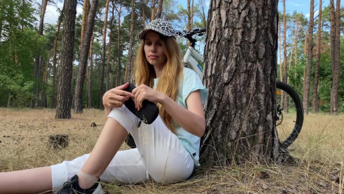 一个戴着棒球帽的女孩在一棵树旁休息，用智能手机看新闻。