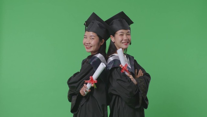 亚洲女学生毕业时戴着帽子，穿着长袍，双臂交叉，对着演播室绿幕背景的镜头微笑