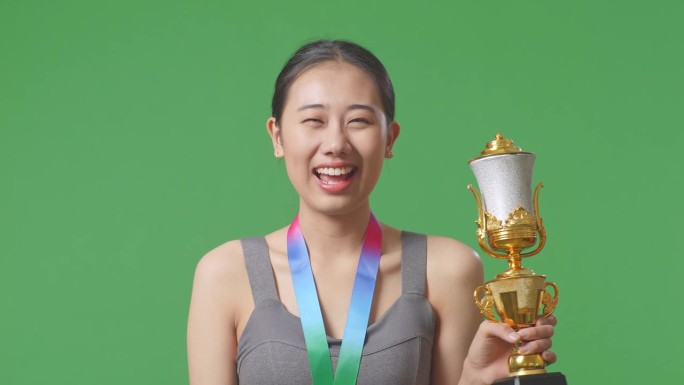 在工作室绿屏背景下，亚洲女子拿着金牌亲吻金杯，自豪地成为第一个获奖者
