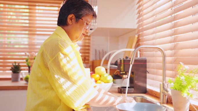 亚洲老年妇女家庭主妇在厨房洗水果。有吸引力的成熟管家清洁和清洗健康的水果和蔬菜用清水准备早餐在家里