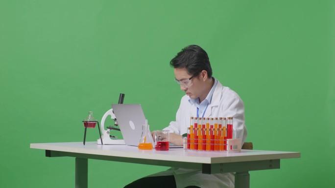 在绿屏背景的实验室里，亚洲男性科学家一边在桌子上用显微镜和试管打字，一边在笔记本电脑上打字
