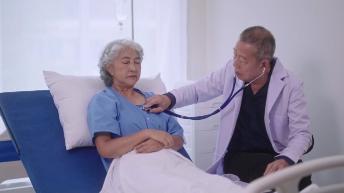 高级亚洲男医生坐在办公桌前，拿着听诊器，听着现代医院里正在康复的老年女病人的心跳、心肺检查。