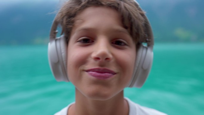 快乐的十几岁的男孩戴着耳机，随着歌曲的节拍动着头。孩子听音乐的特写脸，背景是湖景
