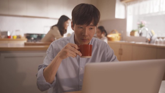 韩国家庭:年轻的父亲在等菜的时候用笔记本电脑远程工作，妻子和孩子在后台准备午餐。支持一个商人的家庭