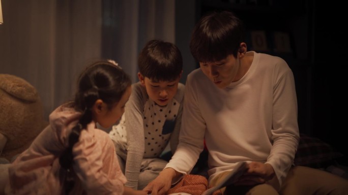 韩国父亲在睡觉前给他可爱的孩子们读童话故事。年轻英俊的爸爸照顾他的孩子，晚上在家读一本书的故事