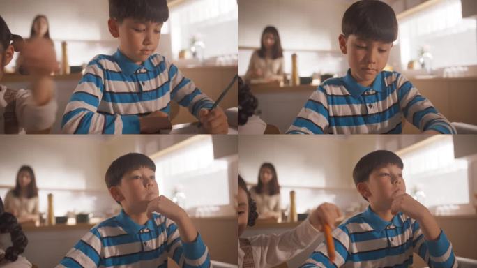 韩国小孩早上坐在家里的餐桌旁的特写照片，背景是妈妈在做饭。聪明男孩思考，做家庭作业。手持特写相机