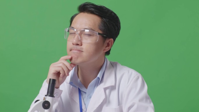 在绿屏背景的实验室里，亚洲男性科学家在显微镜下观察时，思考，环顾四周，举起食指