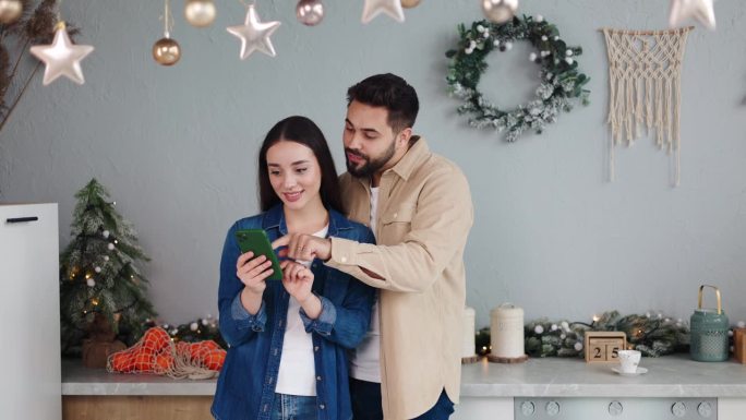 相爱的情侣拿着手机在现代舒适华丽的厨房里享受冬季圣诞新年假期。现代科技的使用，在网上度过空闲时间，在