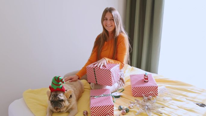 快乐的女人用红盒子包装圣诞礼物，她的狗戴着精灵帽