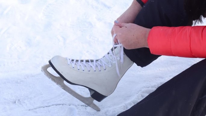 剪短的无法辨认的特写女性双手穿着暖和的夹克，系着冰鞋，系着鞋带，坐在雪地上。冬天的活动