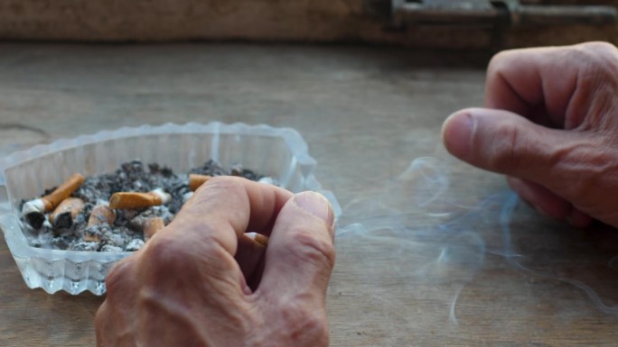 退休老人抽烟。一个老人手握香烟的特写，烟灰缸里有很多烟头。老年人生活中的坏习惯