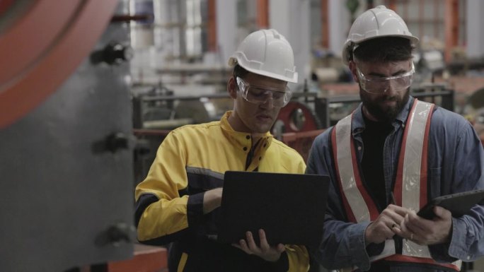 在一家大型工厂里，一队机械厂工程师使用笔记本电脑检查生产钢铁相关部件的机器。专业工作，戴安全帽，保护