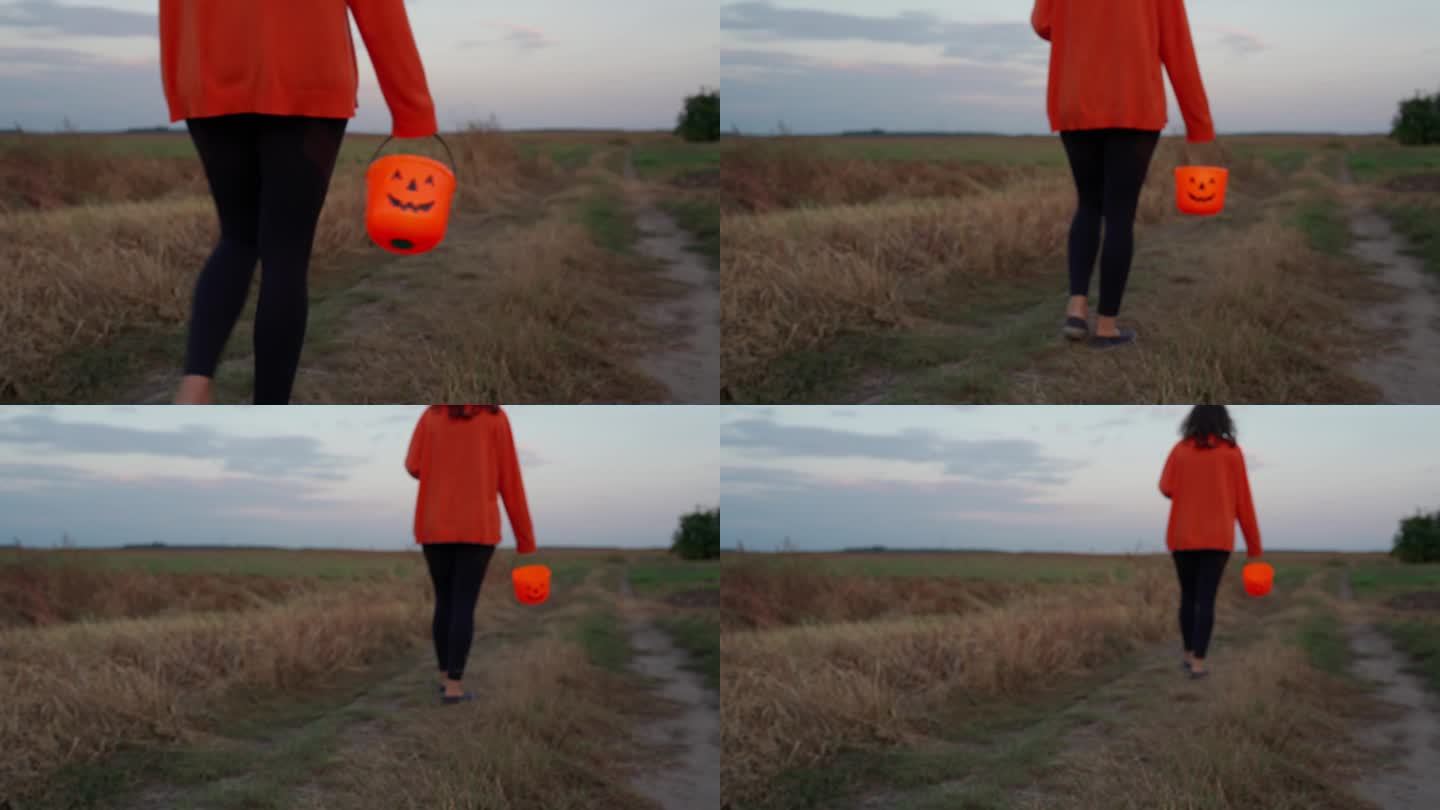 万圣节晚上，一个女孩提着南瓜形的篮子走在田野的路上。不给糖就捣蛋派对的4K概念。在秋天的傍晚，一位年