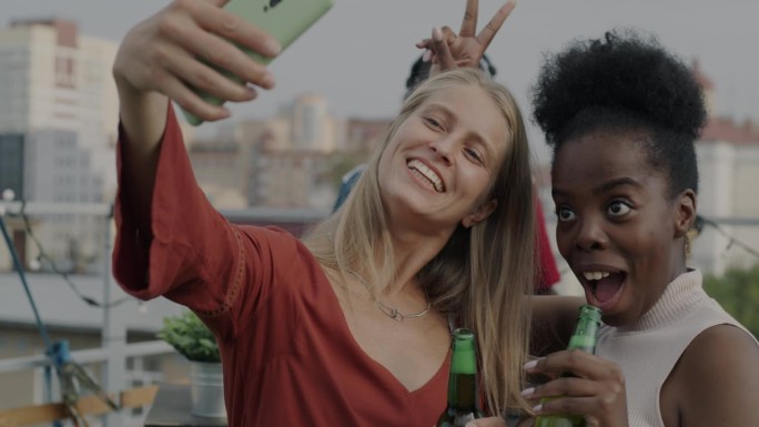 在城市背景下的派对上，年轻的非裔美国人和白人女性拿着饮料对着智能手机自拍