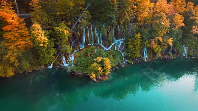 瀑布在色彩缤纷的秋天森林和山湖与清澈的蓝绿色的水在日落。长着黄叶的树。原始的本性。