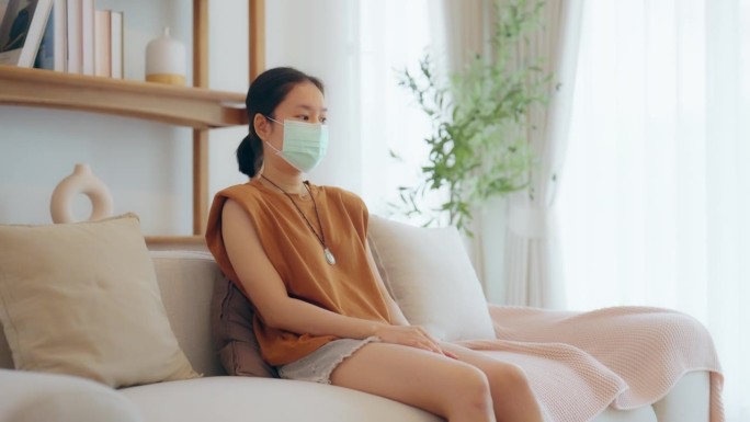 年轻的亚洲妇女坐在沙发上，戴着口罩，感冒发高烧。保健和保险概念