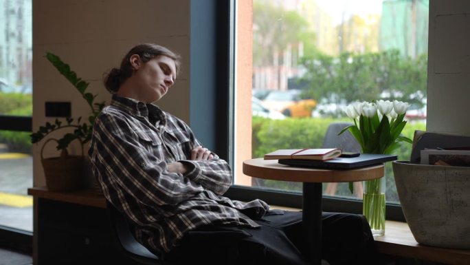 疲倦的年轻人闭着眼睛坐在咖啡馆的桌子旁睡觉。筋疲力尽的白人男子在咖啡馆里小睡，等待点餐。