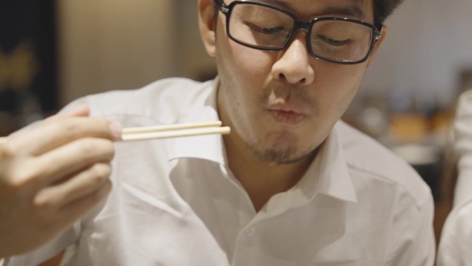一位亚洲男性职员下班后在日本餐厅与同事共进晚餐，脸上带着满意的表情，用筷子吃着蟹肉棒或鱼糜。