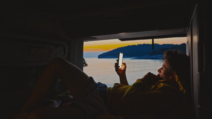 一名男子斜倚在露营车上的床上玩智能手机