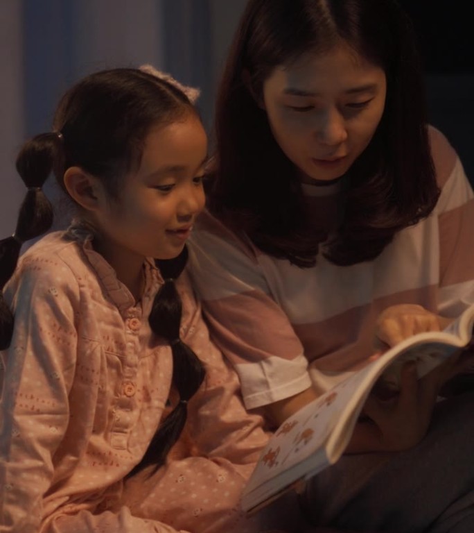 竖屏:韩国母亲临睡前给可爱的女儿读童话故事。年轻美丽的妈妈照顾小女孩，晚上在家读一本书里的故事
