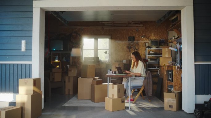 西班牙裔女商人在车库使用笔记本电脑。女独资经营者填写网上客户的订单。产品在纸板箱准备运输。在家工作的