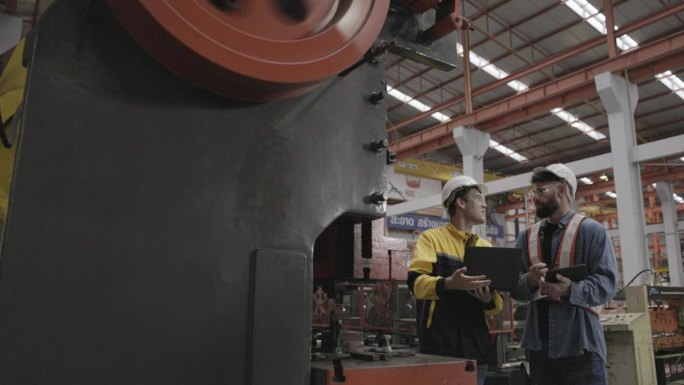 在一家大型工厂里，一队机械厂工程师使用笔记本电脑检查生产钢铁相关部件的机器。专业工作，戴安全帽，保护