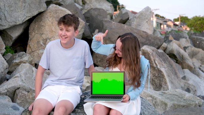 年轻的十几岁的夫妇坐在石头上，用手指指着笔记本电脑，绿色屏幕上有广告文字白色底部蓝色顶部的衣服微笑白