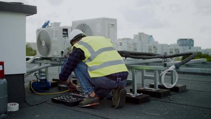 穿着防护装备的男电工在室外屋顶上安装新的空调。安装和修理风扇