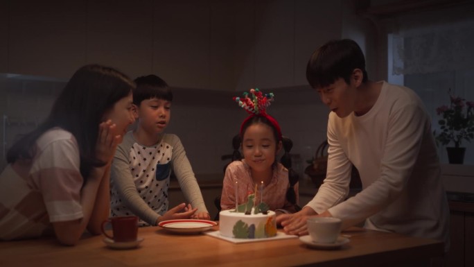 可爱的韩国家庭在晚上用蛋糕和蜡烛庆祝生日。小女孩吹蜡烛，他们鼓掌，笑，开玩笑，一起创造快乐的回忆