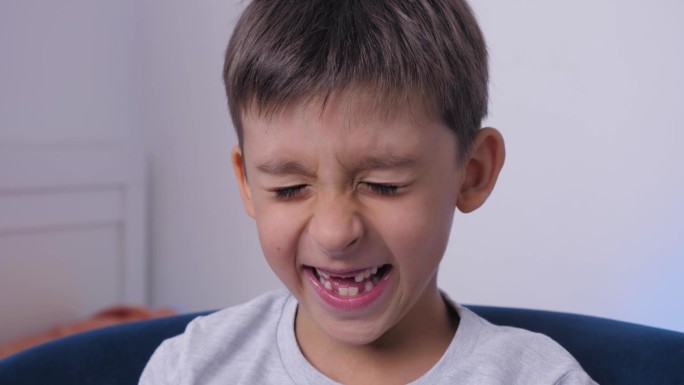 近口小孩掉了一颗乳牙。更换恒换牙、无牙微笑、牙龈破洞。儿童牙科概念。男孩穿着一件灰色t恤，对着镜头哭
