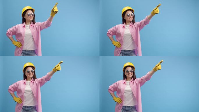 年轻的女工戴着黄色安全帽，戴着美式眼镜和手套。在工作室里做广告，蓝色背景，手指向右制作广告展示标志，
