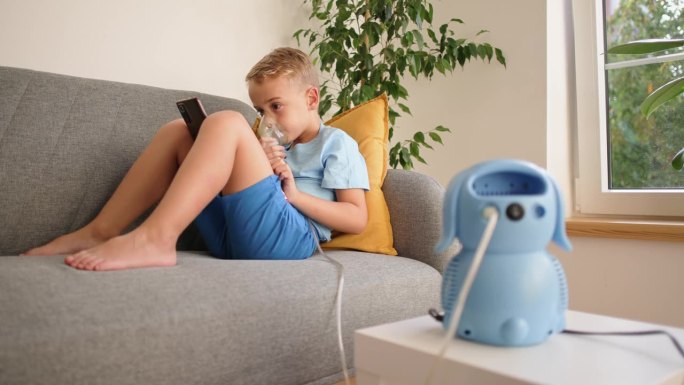 患有哮喘的高加索男孩，在治疗过程中，一边使用雾化口罩，一边坐在沙发上玩手机