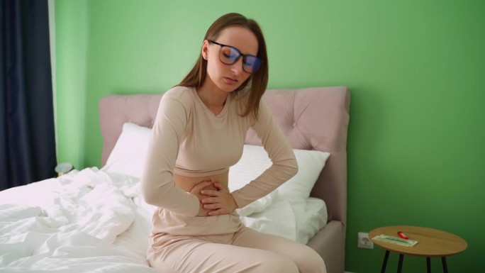戴眼镜的年轻女子，痛经，痛经。一位年轻女子抱着肚子坐在床上，忍受着月经带来的疼痛。
