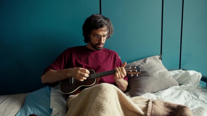 一个留着胡子的黑发男人，穿着红衬衫，戴着眼镜，躺在一张蓝墙的床上弹吉他