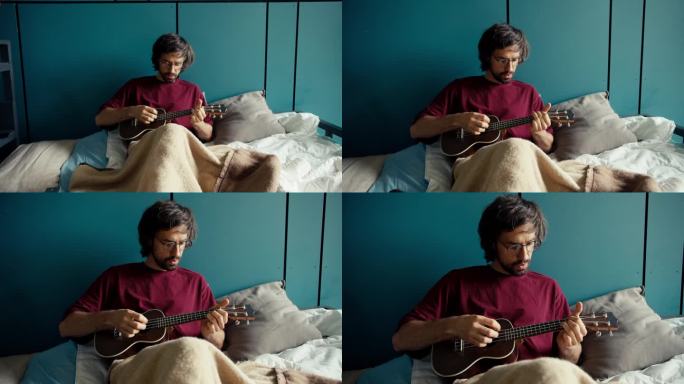 一个留着胡子的黑发男人，穿着红衬衫，戴着眼镜，躺在一张蓝墙的床上弹吉他