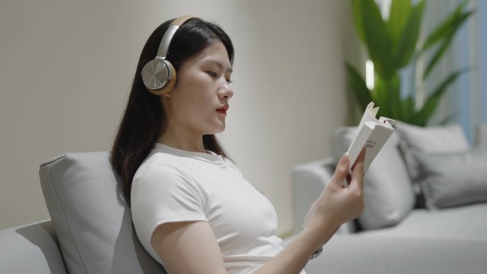 年轻的亚洲女性在沙发上读书，戴着耳机，享受休闲时光