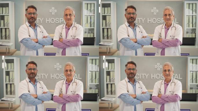 医院中等肖像:两个友好的印度男医生交叉双手，看着镜头微笑。穿着白大褂的成功中年医疗保健医师