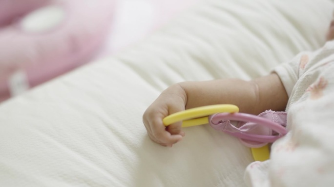 慢镜头:一个刚出生的女婴躺在婴儿床上，抱着并玩着色彩柔和的塑料圈。